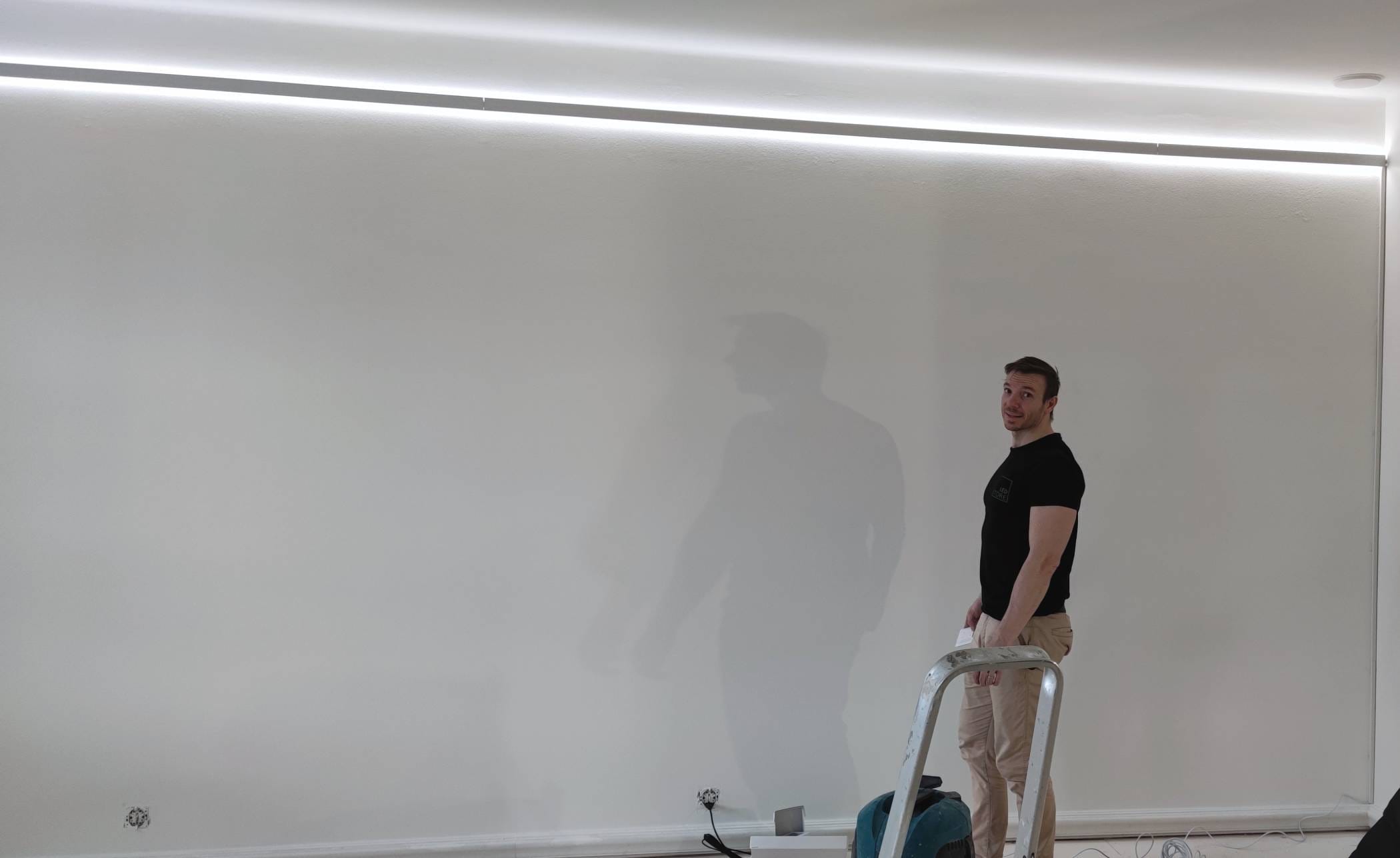 Henkilö tikkailla työkalun kanssa, pehmeässä LED-valaistuksessa yläpuolelta, valkoista seinää vasten. Kaapelit ja laitteet hajallaan lattialla LED-myymäläasennuksissa.